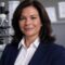Portrait of Dr. Maria Torres, a middle-aged woman with dark brown wavy hair, wearing a navy blazer and white shirt, standing in a forensic lab with a microscope in the background